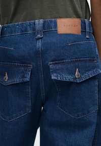 Οπίσθια όψη μπλε τζιν με κουμπωτές τσέπες με καπάκι και καφέ δερμάτινη ετικέτα Topshop στη μέση.