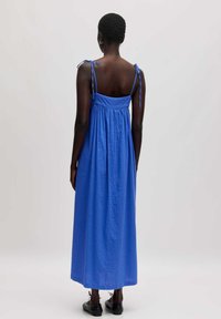 Blaues Maxikleid aus leichtem Stoff mit verstellbaren Bindegurten, gerüschtem Rücken und strukturierter Oberfläche. Die Schlichtheit im Design betont den Komfort.