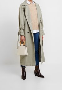 Trench coat verde chiaro con maniche arrotolate, maglione beige, jeans scuri, stivaletti alla caviglia marrone con stampa pitone, e una piccola borsa crema con manici in legno.