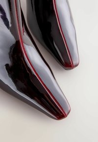 Klæbrige sorte laklæder ankelstøvler med spids tå. Har røde sting langs sømmen og en elegant, glat tekstur.