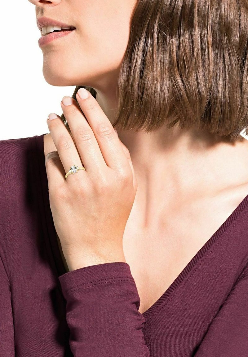 Anillo de oro con una piedra clara central, llevado en una mano que sostiene un pequeño objeto metálico. La persona lleva una blusa de manga larga color burdeos profundo.