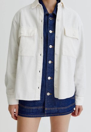 Woman wearing an unbuttoned white corduroy jacket over a buttoned dark blue denim dress with visible pockets and seams.