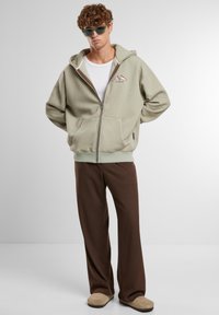 Beżowa bluza z kapturem na zamek, z dużą przednią kieszenią, noszona na białym topie na ramiączkach. Zestawiona z brązowymi spodniami o szerokich nogawkach i beżowymi butami typu slip-on.