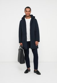 Mann in navy Parka, weißem Shirt, navy Hose und schwarzen Slippern, hält eine schwarze Ledertasche und steht vor einem weißen Hintergrund.