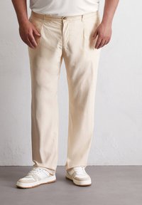 Lichtbeige broeken in een ontspannen pasvorm, met een knoopsluiting, geplooide voorkant en een gladde textuur. Gecombineerd met witte sneakers.