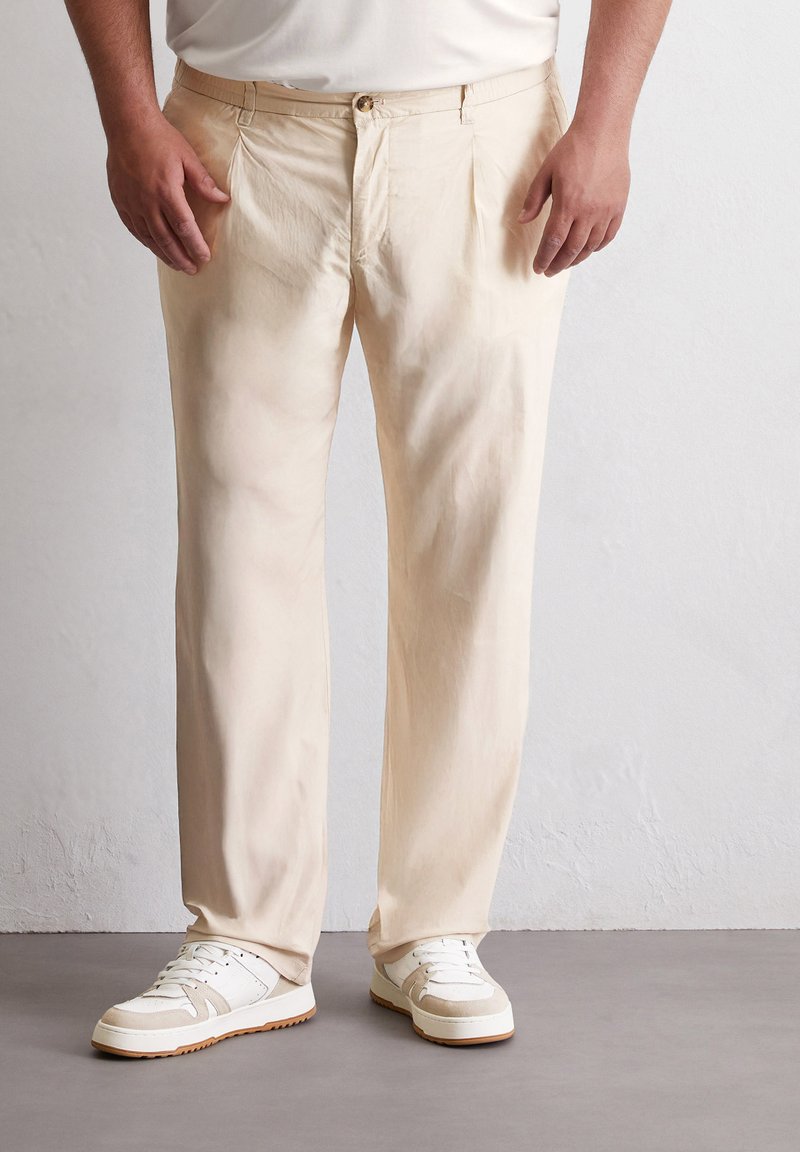 Lichtbeige broeken in een ontspannen pasvorm, met een knoopsluiting, geplooide voorkant en een gladde textuur. Gecombineerd met witte sneakers.