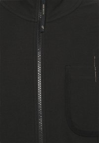 Svart zip-up jacka med texturerad tyg, silverfärgad dragkedja och en bröstficka med kontrasterande sömnadsdetaljer.