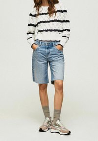 Strickpullover mit Rautenmuster in schwarz-weißen Streifen, kombiniert mit hellblauen Jeansshorts und mehrfarbigen Sneakers mit grauen Socken.