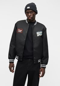 Μαύρο μπουφάν τύπου bomber με λευκές ρίγες στα μανίκια, διακοσμημένο με κεντημένα μπαλώματα "Coors Light" και "Silver Speed", μεταλλικά κουμπιά και υφή με ανάγλυφη επιφάνεια.