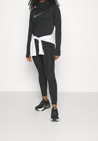 Černý dlouhý rukávový top s vysokým límcem a stříbrným logem Nike, spárovaný s černými legínami a černými teniskami s bílými akcenty. Bílá košile uvázaná v pase.