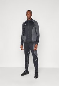 Vyriškas tamsiai pilkas sportinis kostiumas su baltais siuvinėjimo akcentais, apimančiu aukštą apykaklę su užtrauktuku ir siaurėjančiomis kelnėmis, pagamintas iš elastingos medžiagos.