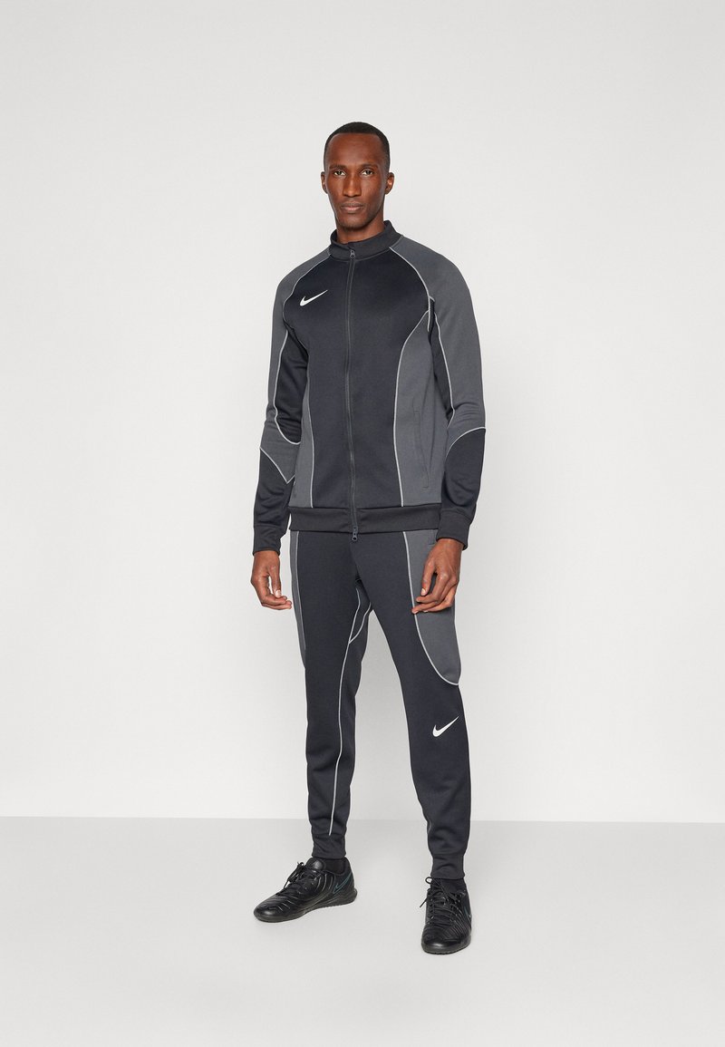 Vyriškas tamsiai pilkas sportinis kostiumas su baltais siuvinėjimo akcentais, apimančiu aukštą apykaklę su užtrauktuku ir siaurėjančiomis kelnėmis, pagamintas iš elastingos medžiagos.