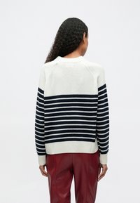 Persona con cabello rizado y largo vista desde atrás, vestida con un suéter blanco con rayas horizontales azul marino y pantalones de cuero rojos.