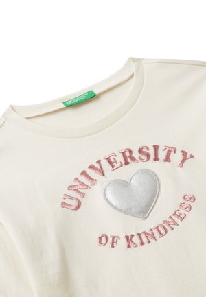 T-shirt color crema con la scritta "UNIVERSITY OF KINDNESS" ricamata in rosa e una grafica a cuore argento su un morbido tessuto di cotone.