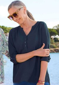 Dunkelblaue Bluse mit Knopfleiste, hochgerollten Ärmeln und glatter Textur, kombiniert mit gemustertem Stoff im Hintergrund.