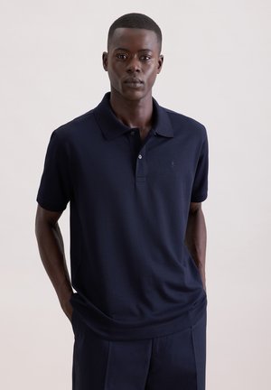 Uomo adulto che indossa una polo a maniche corte blu navy e pantaloni abbinati, in piedi con le mani nelle tasche davanti a uno sfondo chiaro.