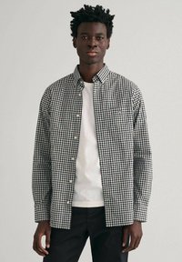 Black and white checked shirt with button-down collar, worn over a white t-shirt, paired with black trousers. Smooth fabric, casual fit.