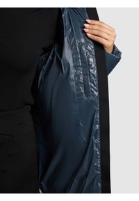 Person klädd i svarta kläder håller upp en mörkblå glansig kappa och visar en innerficka med welt på en ljusgrå bakgrund.