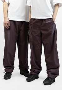Maroon athletic pants with a loose fit, featuring mesh accents along the sides and a drawstring at the cuffs. Paired with black sneakers.