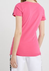Camiseta de algodón rosa de manga corta con cuello redondo y un pequeño logo en la manga. Textura suave, diseño ajustado y aberturas laterales.