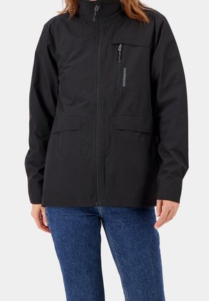 Kobieta ubraną w czarną kurtkę zapinaną na zamek z kieszeniami na klatce piersiowej i na wysokości talii, w połączeniu z niebieskimi dżinsami, stoi na jednolitym tle.