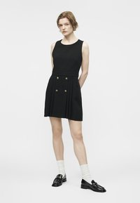 Woman with short hair wearing a sleeveless black dress with front buttons, white socks, and black loafers, standing against a plain background.