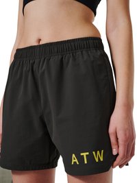 Zbliżenie osoby noszącej czarne szorty z elastycznym pasem z żółtym napisem "ATW" na lewym udzie oraz czarny krótki top.