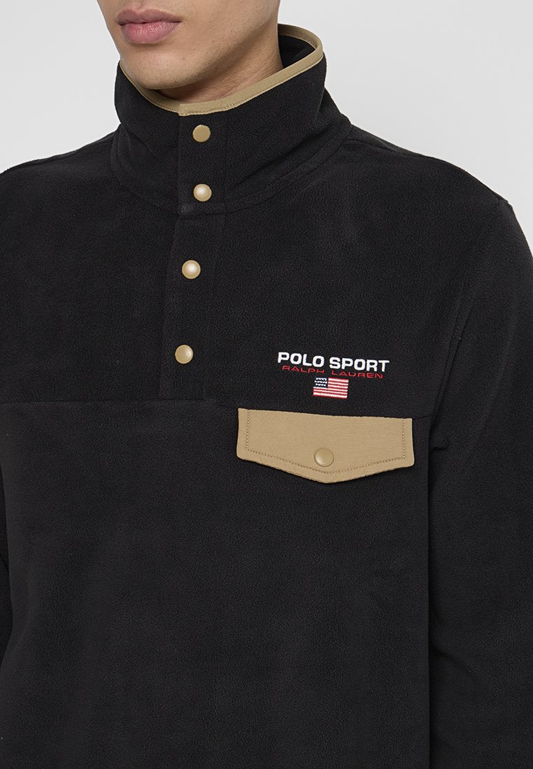 Czarny polarowy golf z beżowymi zatrzaskami, beżową klapką kieszeni i haftowanym logo "Polo Sport Ralph Lauren" z amerykańską flagą.