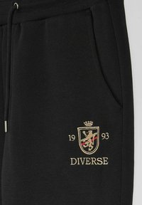 Czarne dresy wykonane z miękkiego materiału, z haftowanym złotym logo przedstawiającym koronę, lwa oraz słowo "DIVERSE" z rokiem 1993.
