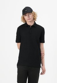 Svart polo-skjorte med teksturert mønster, korte ermer og knapper i kragen. Kombinert med en svart caps med logo og mørke bukser.