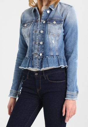 Femme portant une veste en jean bleue cintrée avec un ourlet à volants et des boutons blancs, associée à un jean bleu foncé sur un fond uni.