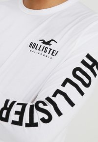 Vit långärmad t-shirt i bomull, med en svart logotyp på bröstet och stor svart text som säger "HOLLISTER" längs ärmen.
