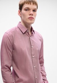 Camicia a maniche lunghe con motivo rosso, dotata di colletto e con una sottile trama. Presenta bottoni bianchi lungo la parte frontale. Il modello posa con un'espressione neutra.