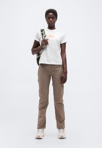 Άτομο όρθιο που φοράει μπεζ παντελόνι, άσπρο μπλουζάκι με σχέδιο, ανοιχτόχρωμα παπούτσια πεζοπορίας και κρατάει ένα πράσινο στρωματάκι τυλιγμένο πάνω σε έναν ώμο.