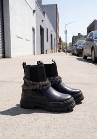 Pair of black chunky sole ankle boots with elastic sides and silver bead chain embellishments on a city sidewalk.