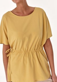 Top amarillo de manga corta con escote amplio y cintura fruncida, que incluye un cordón para un ajuste regulable, fabricado en una tela suave y ligera.