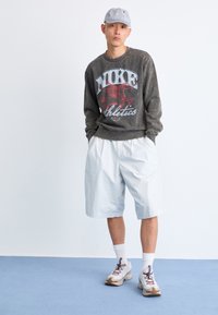 Grå sweatshirt med rød "Nike Athletics" grafik, lyseblå shorts, hvide sneakers og en stribet cap. Casual atletisk stil.