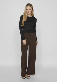 Zwarte top met lange mouwen, gecombineerd met bruine wijde broek met een koord in de taille. Model draagt zwarte sandaaltjes met banden. Eenvoudig ontwerp.