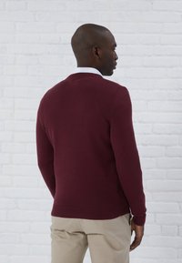 Pulover burgund cu guler și margini texturate, dintr-un material tricotat, purtat peste o cămașă albă cu guler, asortat cu pantaloni bej.