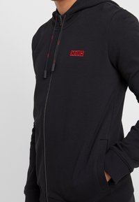 Felpa nera con zip realizzata in misto cotone. Presenta un piccolo logo ricamato rosso, cappuccio regolabile con cordino e tasche laterali.