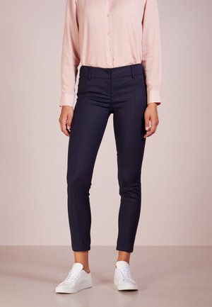 Persona con pantalones ajustados azul marino, camisa rosa pálido abotonada y zapatillas blancas, de pie contra un fondo liso y claro.