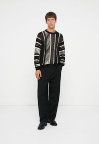 Sort og beige strikket sweater med geometriske mønstre, rund halsudskæring og ribbede manchetter, parret med sorte brede bukser og sorte sko.