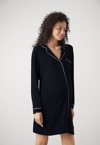Svart langermet nattskjorte med knapper foran, hvit piping på kragen og ermene, og en lomme på venstre bryst. Mykt stoff, tettsittende design.