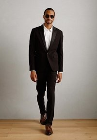 Hombre con traje negro, camisa blanca y zapatos de vestir marrones caminando frente a una pared gris lisa, usando gafas de sol oscuras.