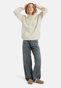 Beige hooded sweatshirt met een voorzak, gecombineerd met grijze wijde jeans en tan instapschoenen. De stof lijkt zacht en casual.