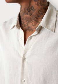 Hellbeige Leinenhemd mit Kragen, zwei Knöpfen und einem strukturierten Stoff. Sichtbare Nähte und eine leicht lockere Passform.