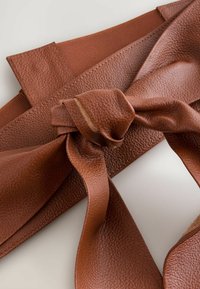 Brun läderaccessoar med en strukturerad yta, som har en knuten rosett och ett elastiskt band i ena änden, vilket betonar dess mjuka flexibilitet.