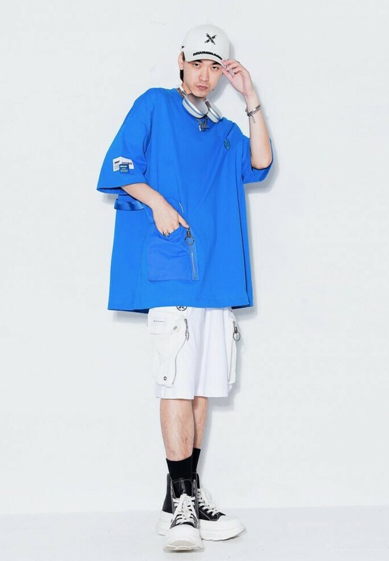 Camiseta azul de gran tamaño con mangas cortas y un bolsillo frontal, combinada con pantalones cortos cargo blancos que cuentan con varios bolsillos y cremalleras, y zapatillas negras y blancas.