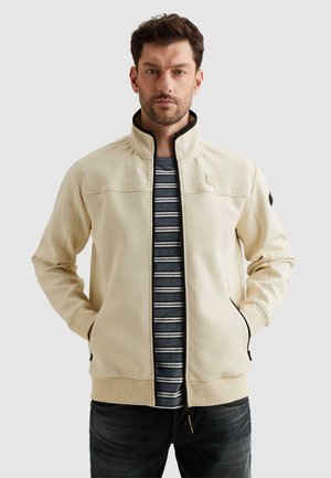 Man wearing a beige zip-up jacket over a black and white striped shirt with hands in jacket pockets, against a plain background.