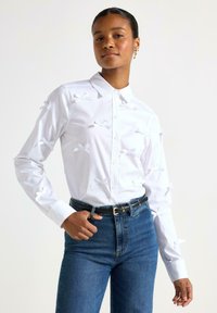 Camicia bianca con bottoni e fiocchi di tessuto sulle maniche, abbinata a jeans blu a vita alta. La camicia ha un colletto classico e chiusura a bottoni.